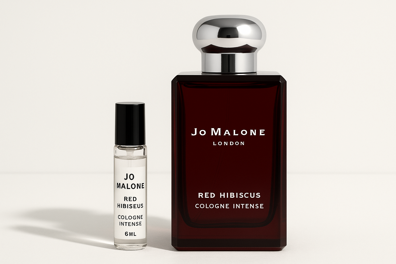 Jo Malone Red Hibiscus Cologne Intense דוגמית 6 מ"ל עומדת ליד בקבוק בושם 100 מ"ל של Jo Malone Red Hibiscus Cologne Intense עם כתובה לבנה על בקבוק אדום כהה
